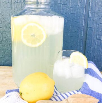 A glass pitcher of fresh squeed lemonade stand behind a glass of lemonade and a yellow lemon