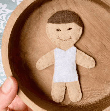 handmade felt doll child wearing a white bodysuit lay in a wooden bowl held over a blue and white tablecloth